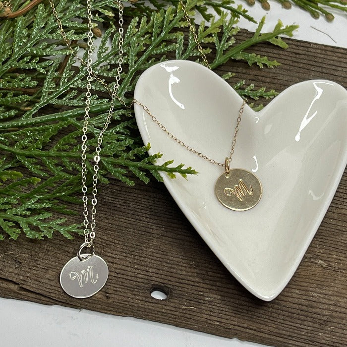 Sterling silver and 14K Gold-filledScript Initial M necklace laying on a heart shaped dish .  Select the length of chain you would like as well as the metal type.
