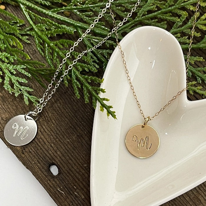Sterling silver and 14K Gold-filled hand-stamped Script Initial M necklaces laying on a heart shaped dish .  Select the length of chain you would like as well as the metal type.